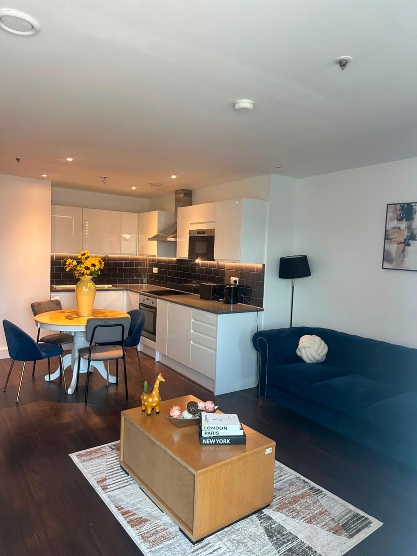 Een moderne woonkamer met een bank, een bijzettafeltje met boeken, een eethoek met stoelen en een vaas, en een keuken op de achtergrond.