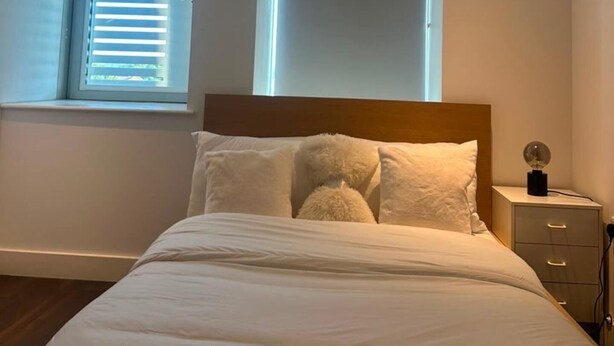A bedroom with a bed, a nightstand, a lamp, and a window with blinds. - London Bliss Apartment - Cozy Stay With City Views (London)