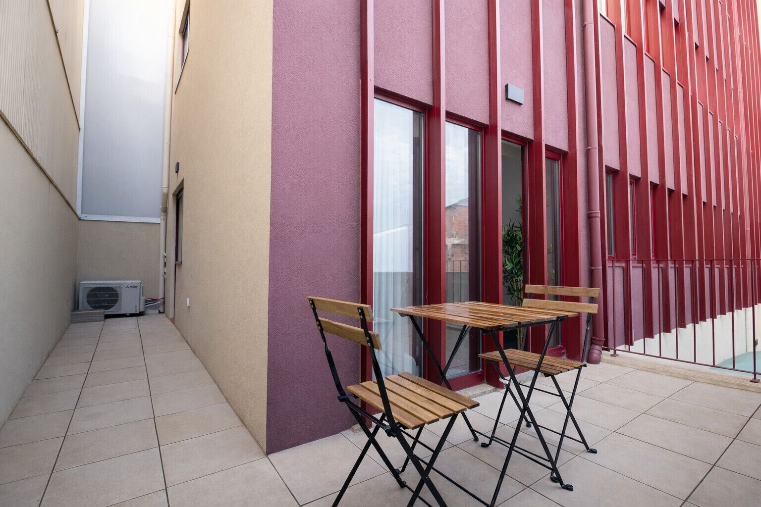 Un espacio al aire libre con dos mesas de madera y sillas, fachada de un edificio rojo y una pizarra a la izquierda.