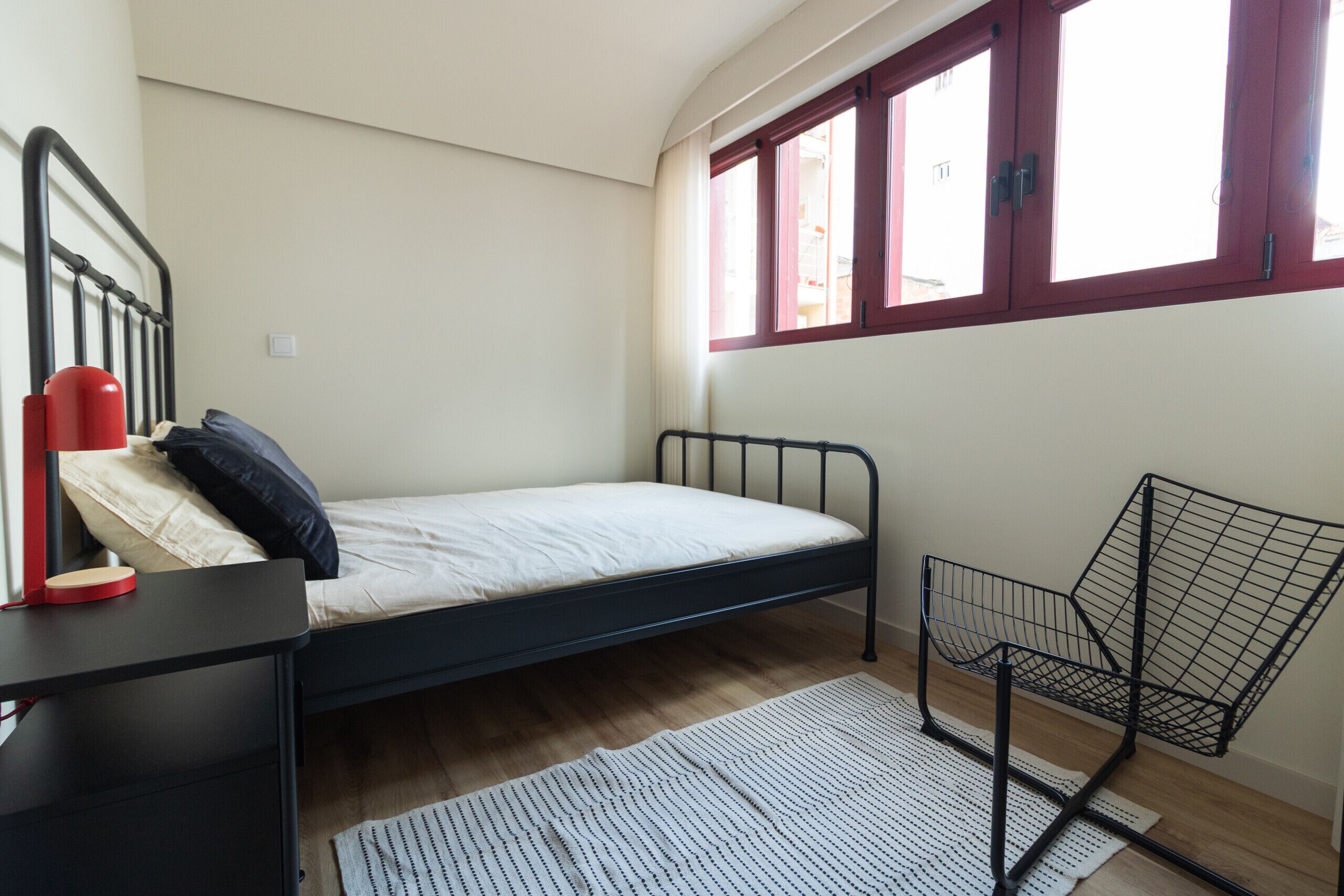 A single bed with a metal frame, a small bedside table with a red object, a chair, and a window with red frames.
