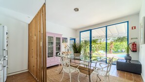 A modern kitchen with a glass dining table, white chairs, a pink cabinet, and a view of a garden.