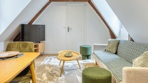 Une pièce de style « loft » avec un plafond en pente, un téléviseur posé sur un meuble en bois, un fauteuil vert rembourré, un canapé à motifs et une petite table ronde.