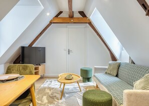 Ein Loft-Zimmer mit schrägem Dach, einem Fernseher auf einem Holzbodenständer, einem grünen gepolsterten Sessel, einer gemusterten Couch und einem kleinen runden Tisch.