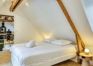 A bedroom with a sloped ceiling, a bed with white linens, a bedside table with a lamp, and a view of a kitchen area. - Bienvenue à L’Olive, votre écrin vert nature au cœur de Rouen. (Rouen)