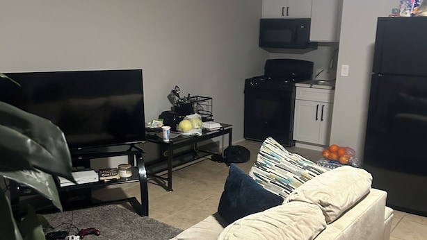 Ein Wohnzimmer mit Sofa, Fernseher, Küchenzeile mit Schränken und Kühlschrank sowie einem Esstisch mit einigen Gegenständen darauf.