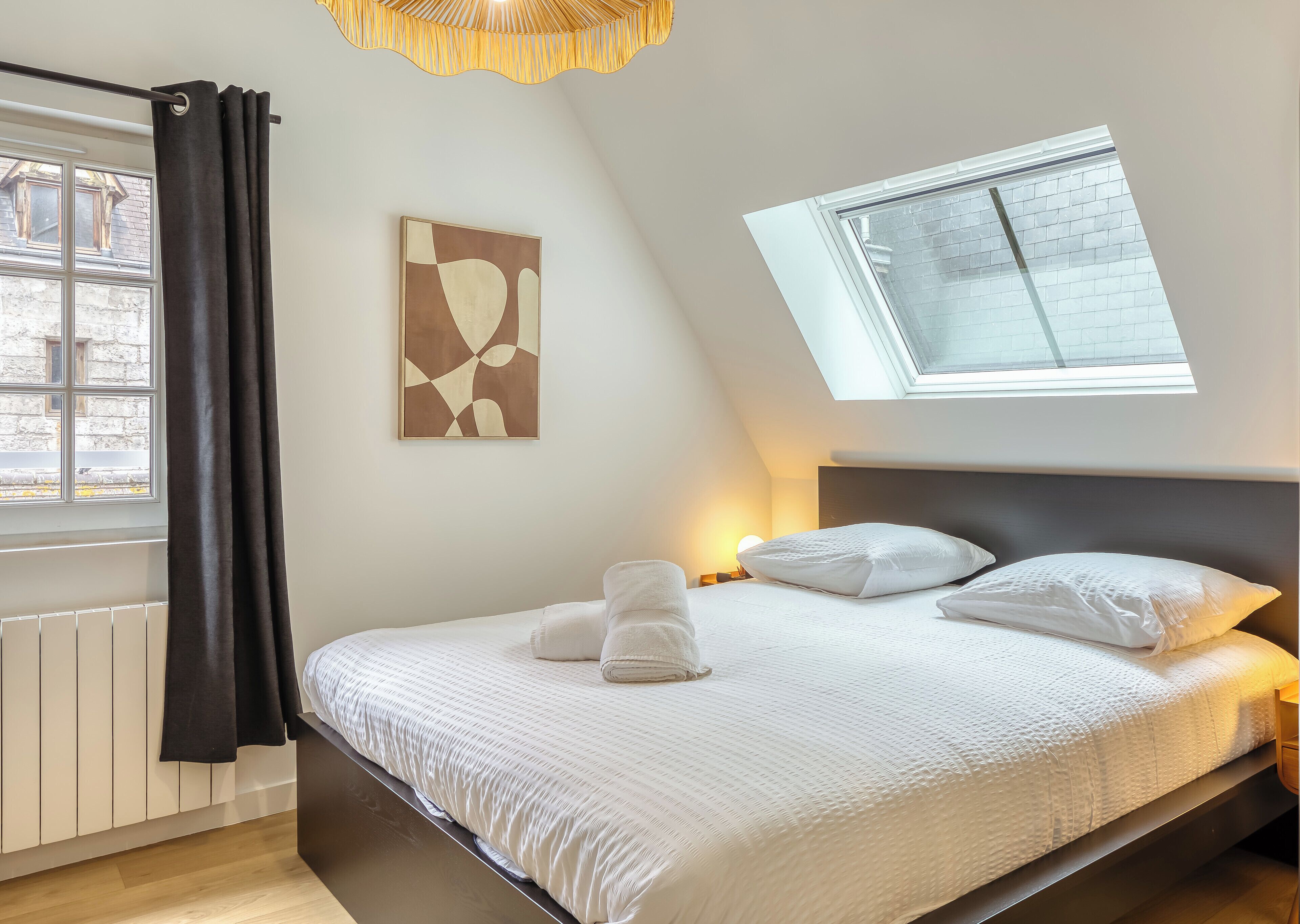 Un dormitorio con cama, un tragaluz, una ventana, un radiador y una obra de arte colgada en la pared.