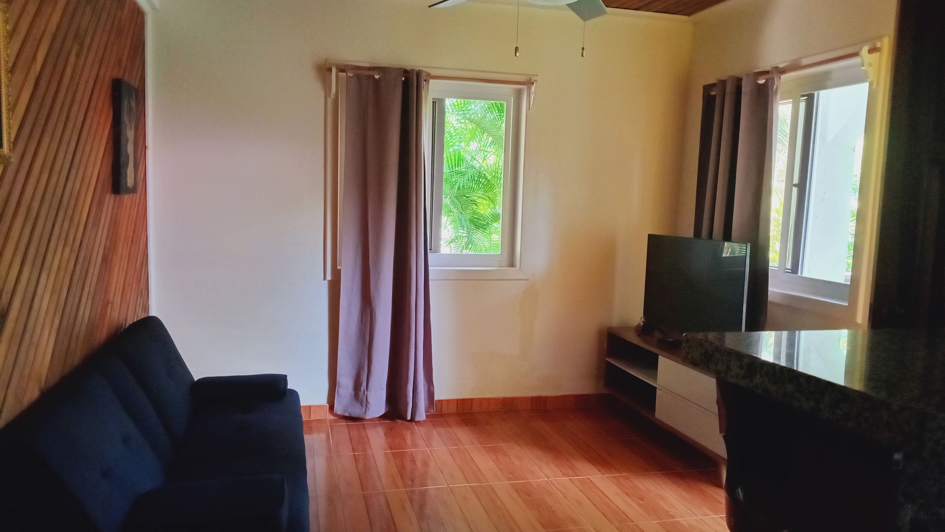 Quarto com piso de madeira, televisão sobre suporte, janela com cortinas e prateleira fixada na parede.