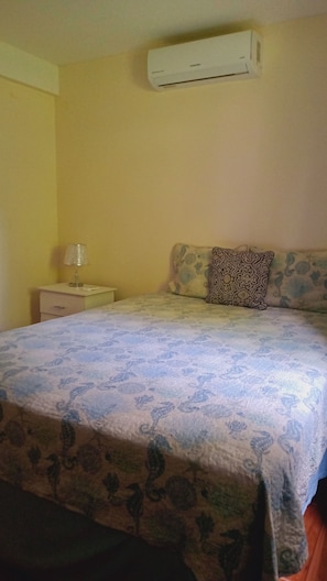 A bedroom with a bed, a nightstand, a lamp, a patterned pillow, and an air conditioner on the wall.