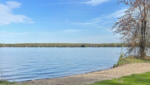 Uma área gramada à beira de um lago, com céu claro e uma linha de árvores ao longe.