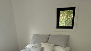 Uma cama bem arrumada com edredom branco e travesseiros, uma pequena janela com vista para árvores e piso de madeira.