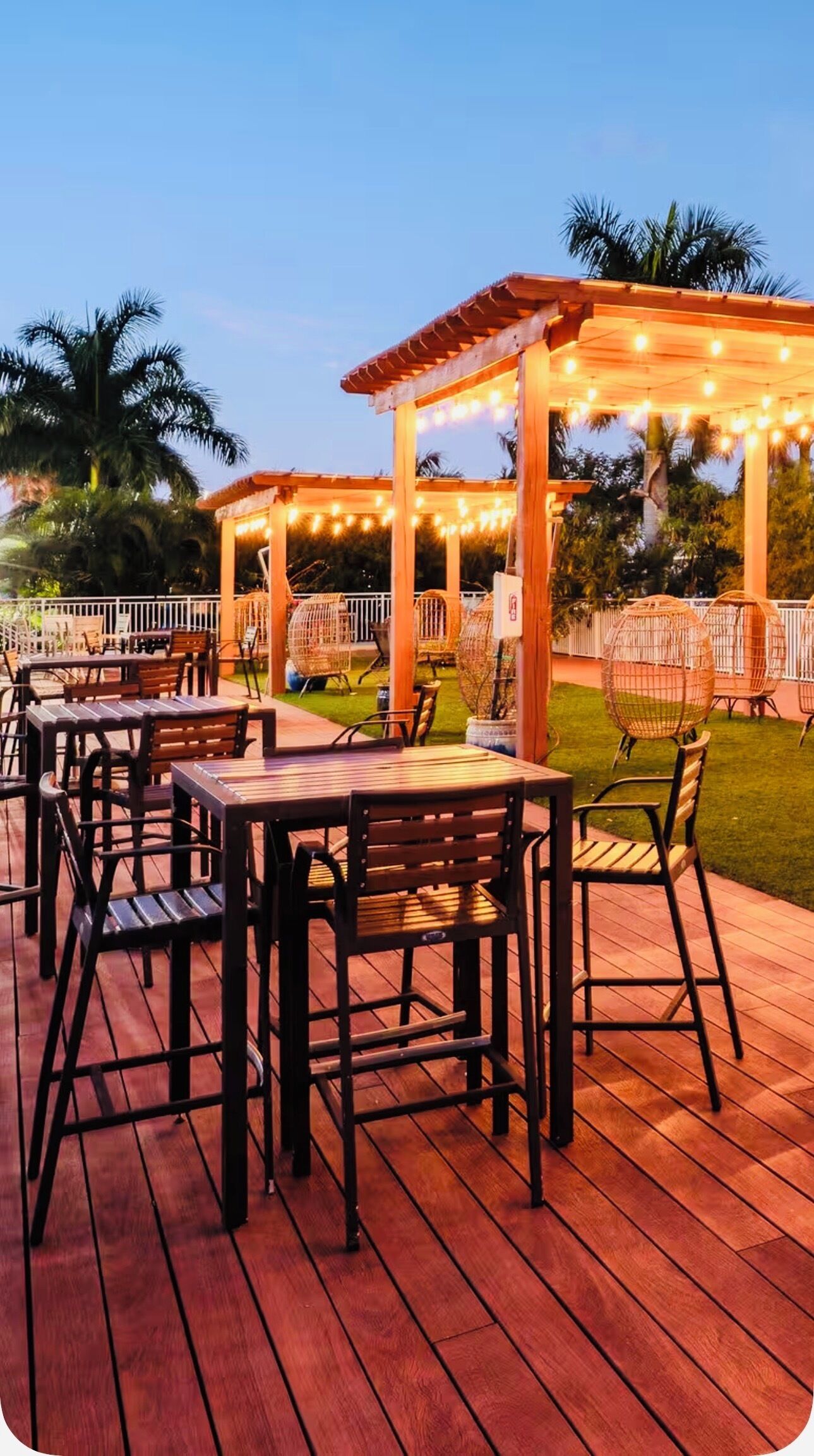 Een terras buiten met houten tafels en stoelen, een pergola met lichtslingers en palmbomen op de achtergrond.