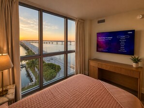 A hotel room with a large window, a bed, a flat-screen TV, and a bedside table with a lamp. - Lake views- Luxury 2BR-2BA - Pool - Walk to Downtown (Fort Myers)