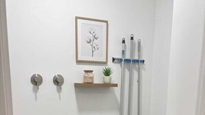 Un lavadero pequeño con lavadora de carga frontal, un estante con una planta y un cuadro en la pared.