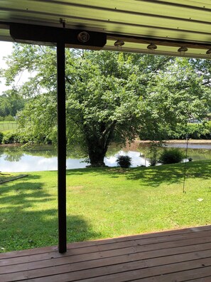 Ein überdachter Außenbereich mit Holzterrasse, einer Metallpergola und Blick auf einen Teich mit Bäumen.