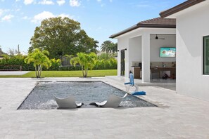 A modern outdoor pool with a unique wave-like design, surrounded by a well-maintained garden and a white building with a visible kitchen area. - El Castillito (Miami)