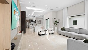 Un salon moderne avec un canapé, des fauteuils, un téléviseur et une cuisine en arrière-plan.