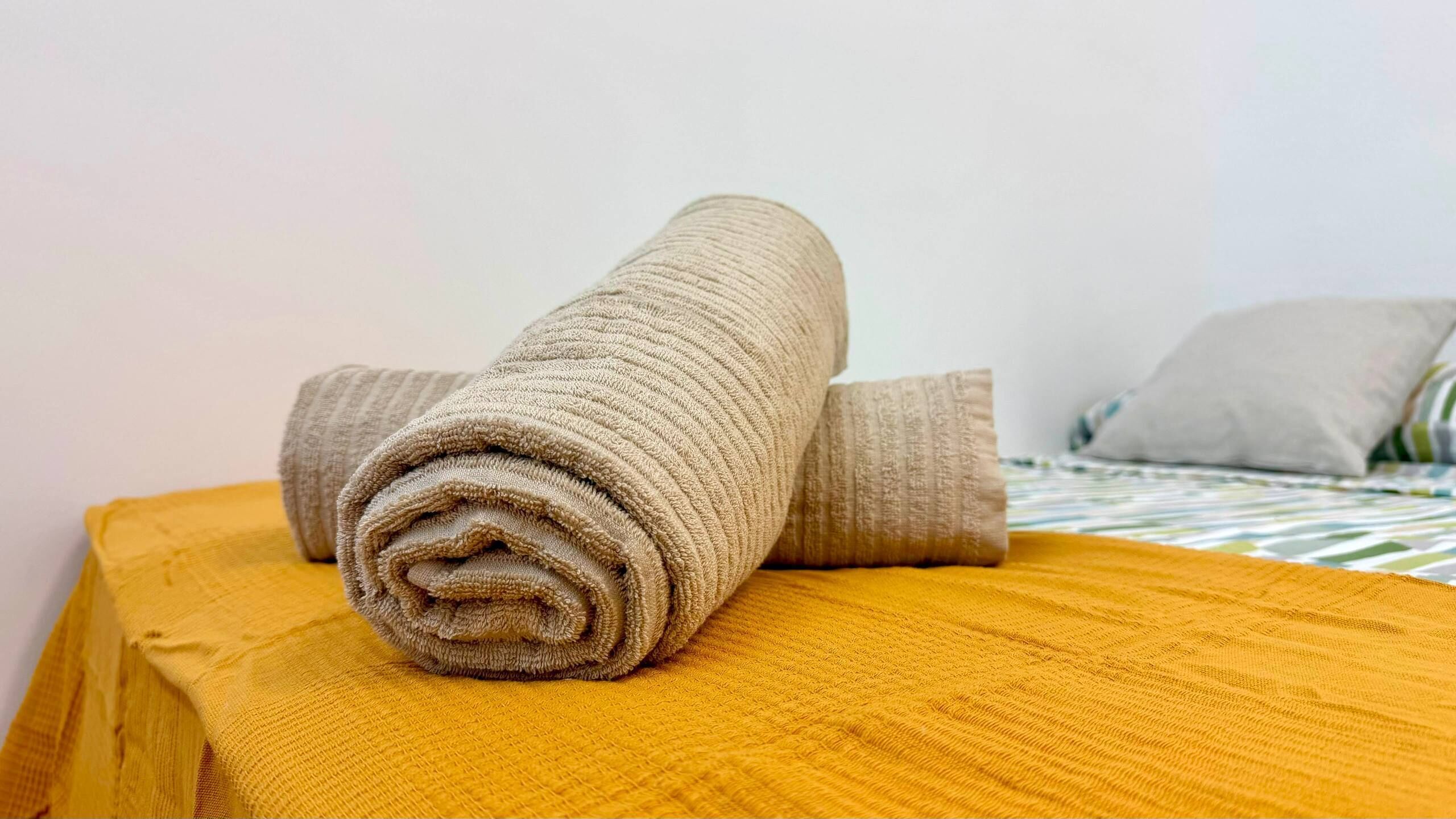 A neatly folded beige towel on a wooden surface, a bed with a striped bedspread, and a pillow in the background.