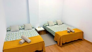 Dos camas individuales con cubrecamas amarillos, una mesita de noche pequeña y blanca, y un tapete gris sobre un piso de madera en una habitación con paredes blancas.