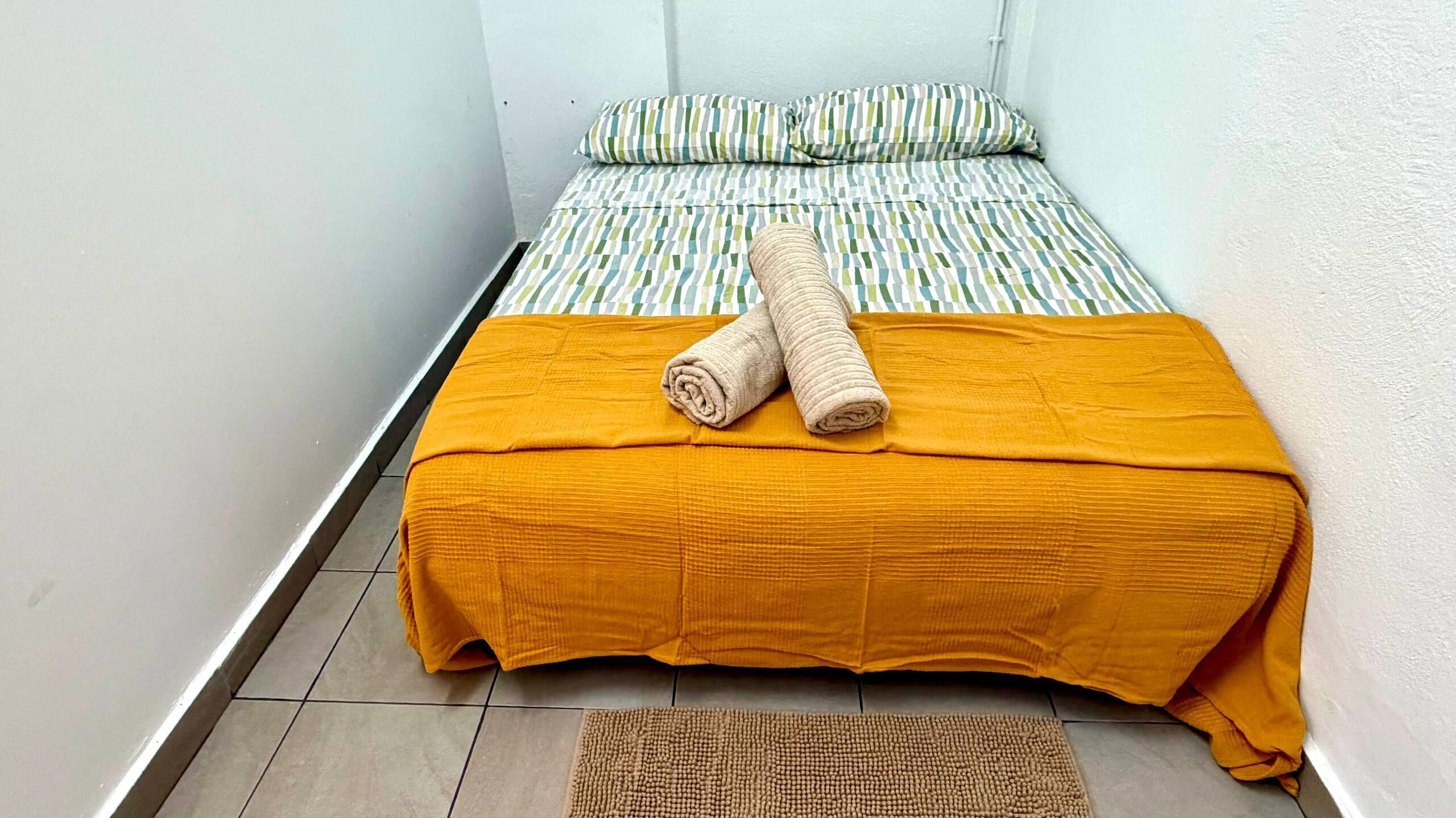 Una habitación pequeña y estrecha con una cama, una toalla enrollada y un tapete.