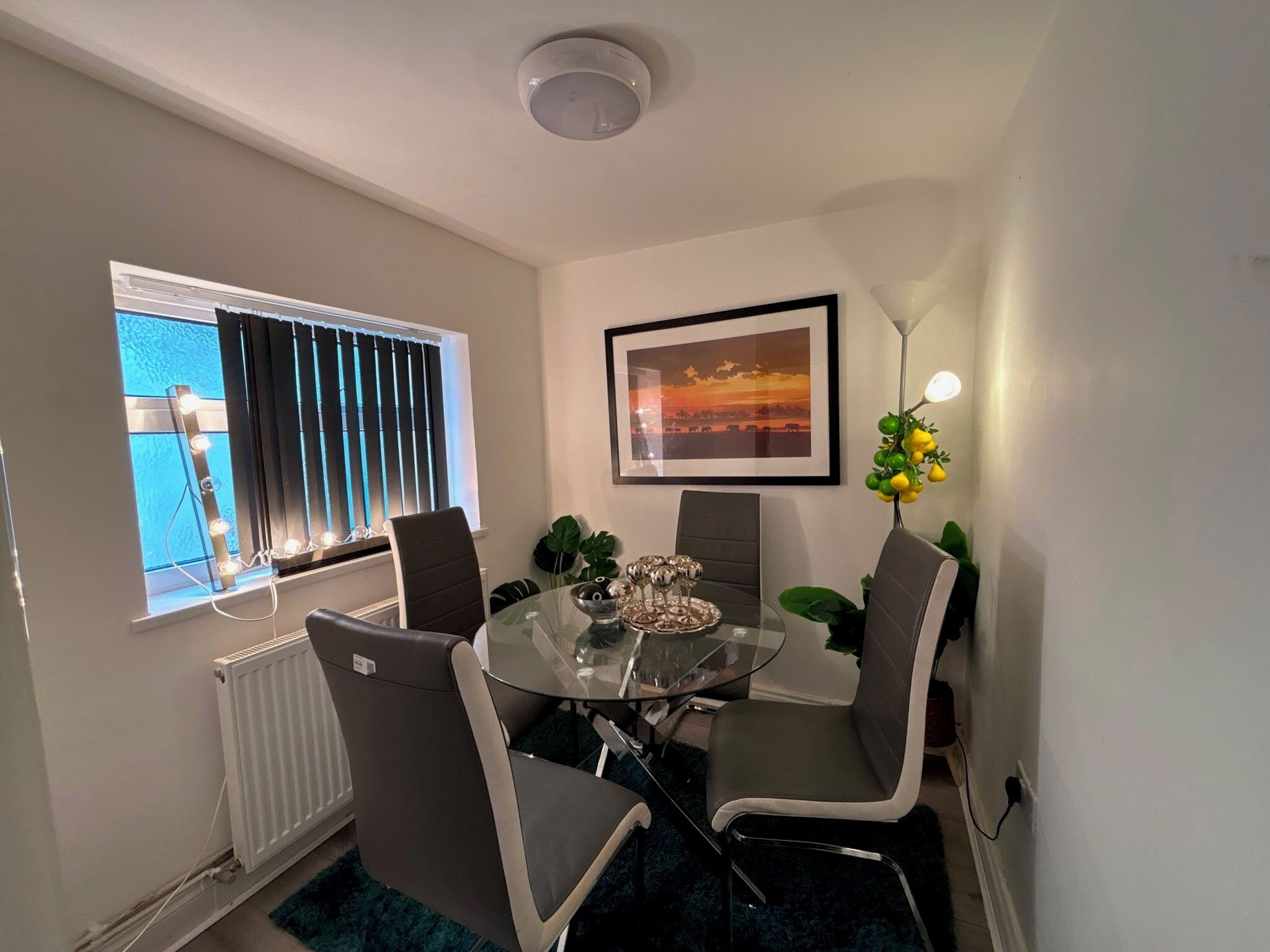 A dining area with a glass table, four chairs, a framed picture, a radiator, and a window with blinds.