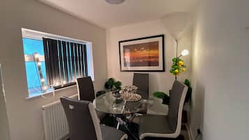 A dining area with a glass table, four chairs, a framed picture, a radiator, and a window with blinds.