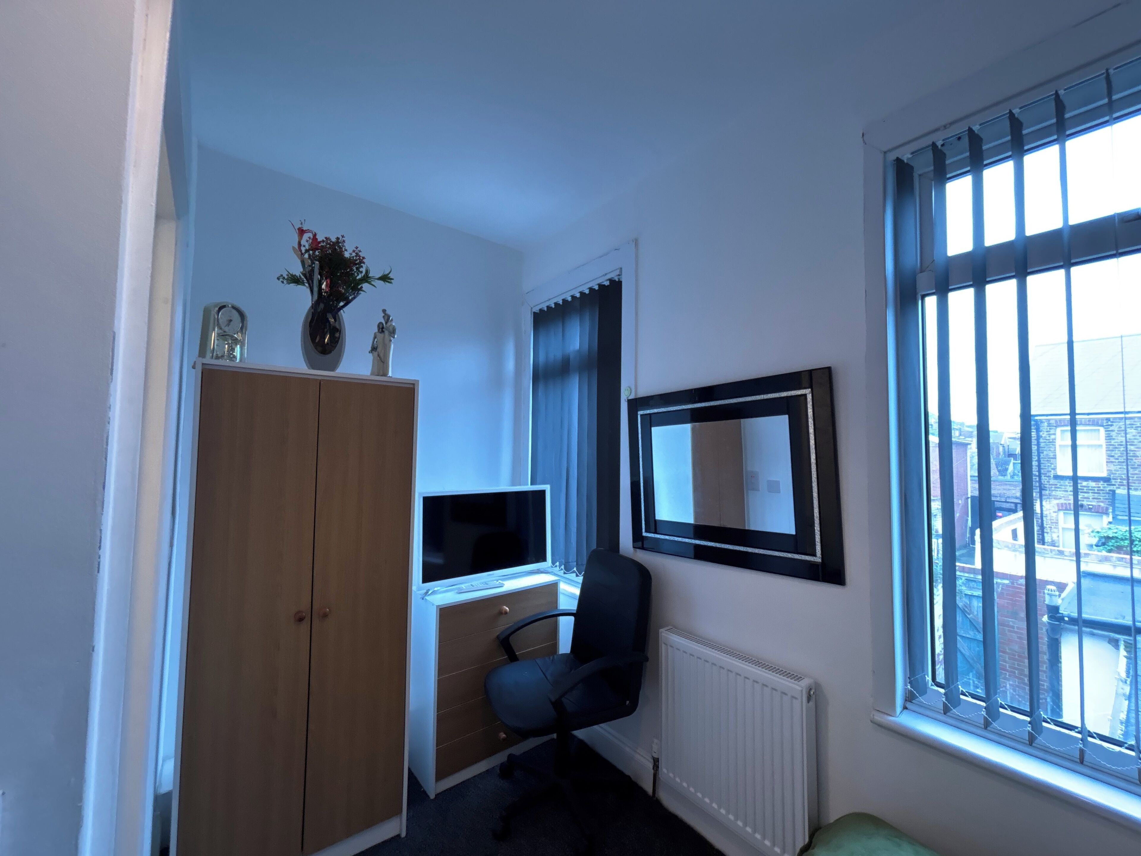 A room with a wooden wardrobe, a desk with a computer, a chair, a radiator, and a window with bars.