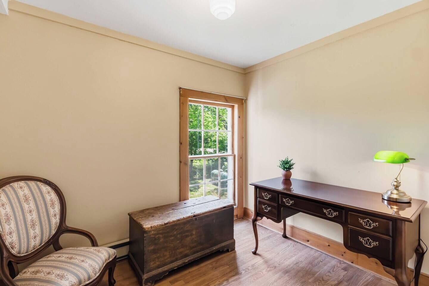 A room with a wooden desk, a chair, a small table with a lamp, and a window with a view of greenery.