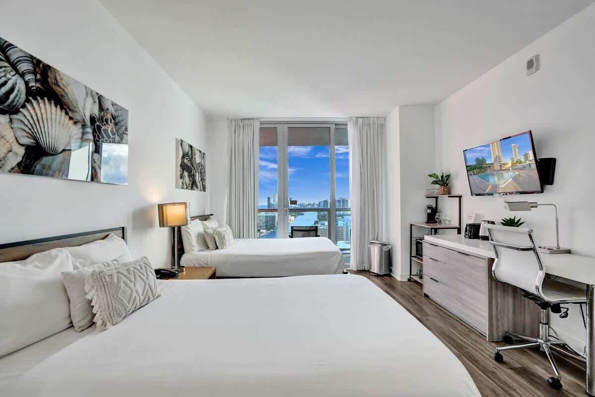 A hotel room with two beds, a desk, a TV, and a large window with a city view.