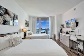 A hotel room with two beds, a desk, a TV, and a large window with a city view. - Modern 2BR Apartment | Exclusive Resort 2609 (Hallandale Beach)