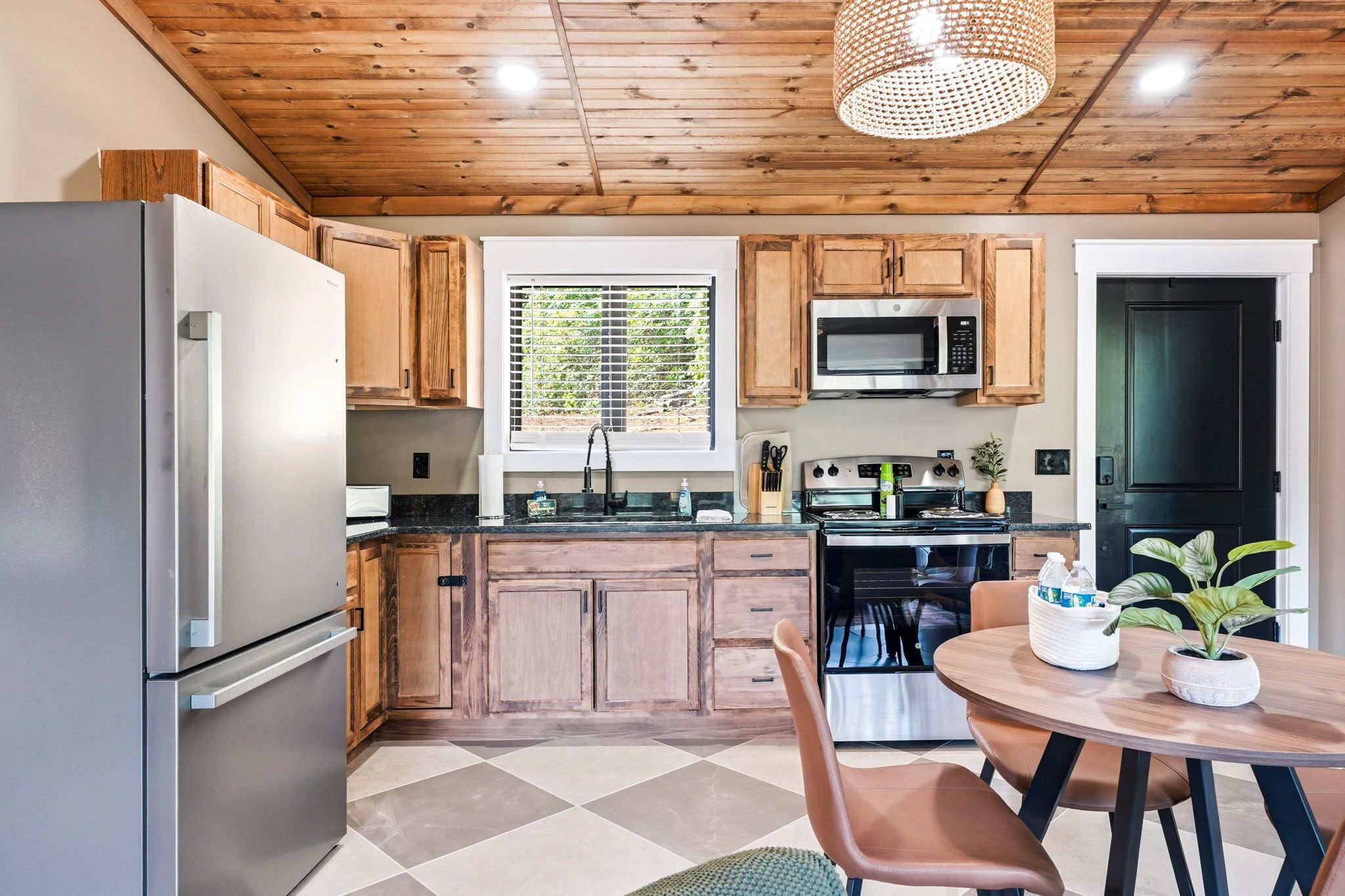 A modern kitchen with wooden cabinets, a stainless steel refrigerator, a microwave, and an oven. There is a window with blinds, a dining table with chairs, and a hanging light fixture.