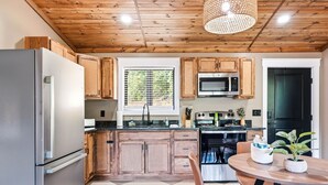 A modern kitchen with wooden cabinets, a stainless steel refrigerator, a microwave, and an oven. There is a window with blinds, a dining table with chairs, and a hanging light fixture. - Cozy 1 bedroom cabin with a hot tub in the mountains of Elizabethton (Elizabethton)