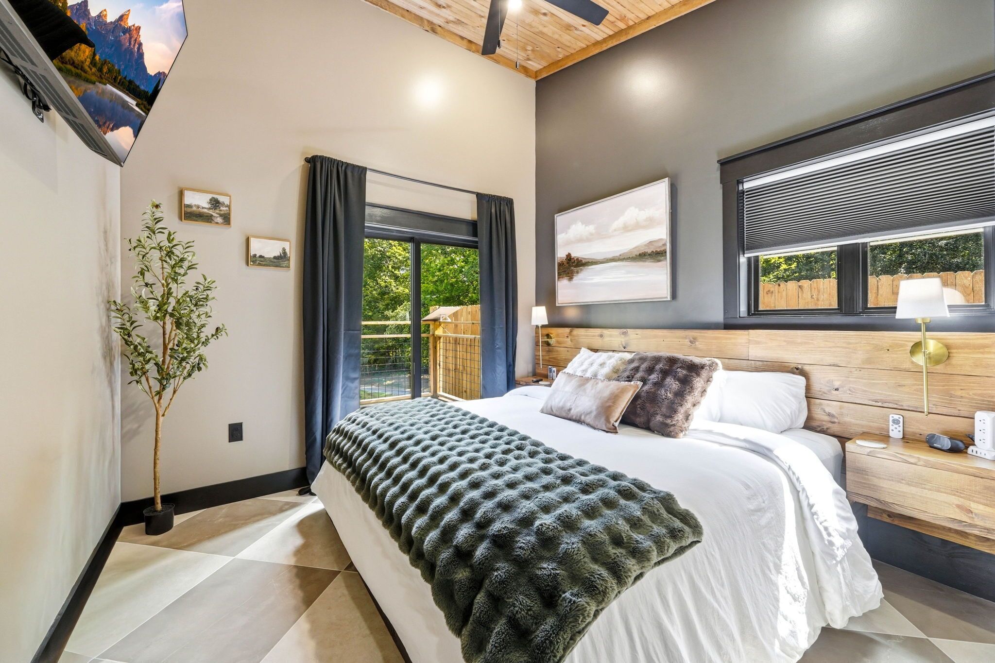 A bedroom with a bed, a potted plant, a window with curtains, a wall-mounted TV, and a painting.