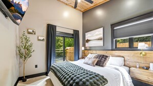 A bedroom with a bed, a potted plant, a window with curtains, a wall-mounted TV, and a painting. - Cozy 1 bedroom cabin with a hot tub in the mountains of Elizabethton (Elizabethton)