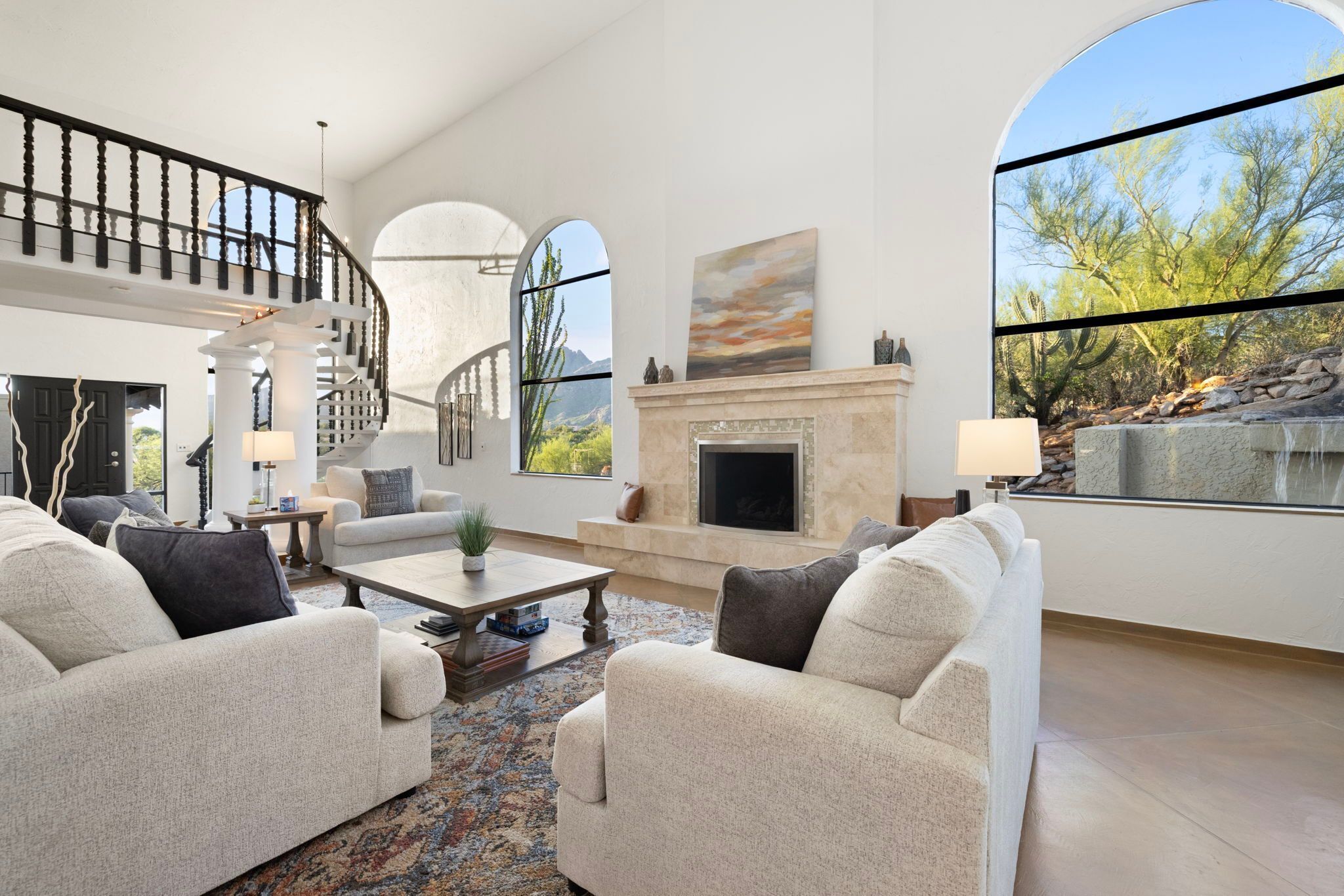 A spacious living room with a fireplace, a staircase, and large windows offering a view of the outdoors.