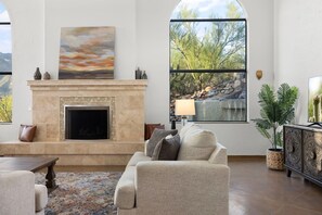 A living room with a fireplace, a sofa, a coffee table, and a large window with a view of trees and rocks. - Sundance  Resort Style Home in the Foothills (Tucson)