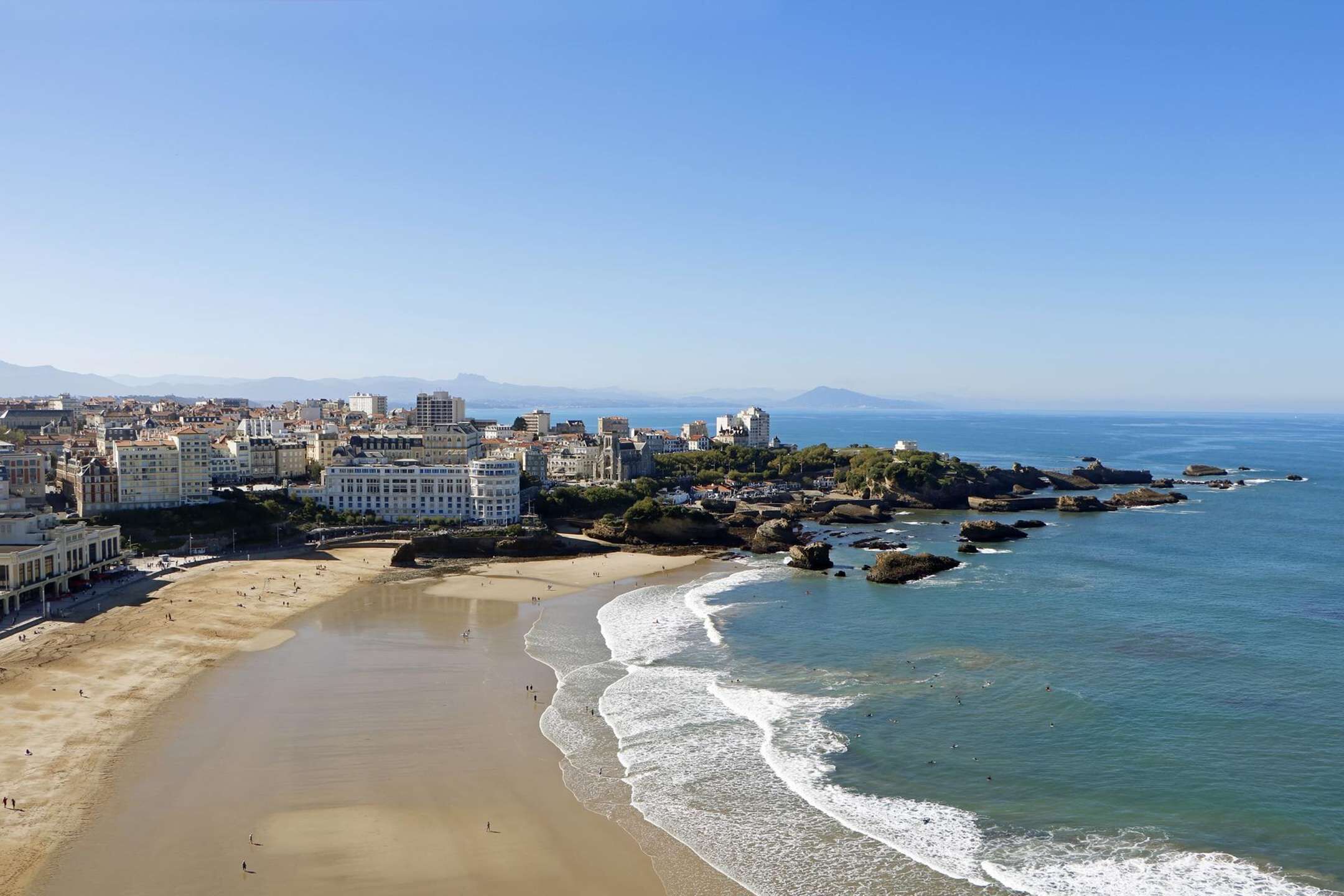 A coastal cityscape with a sandy beach, clear blue sea, and buildings along the shore.