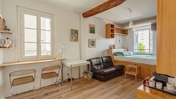 Une pièce de taille modeste, comprenant un bureau, deux chaises, un canapé, un lit et un petit tabouret.
