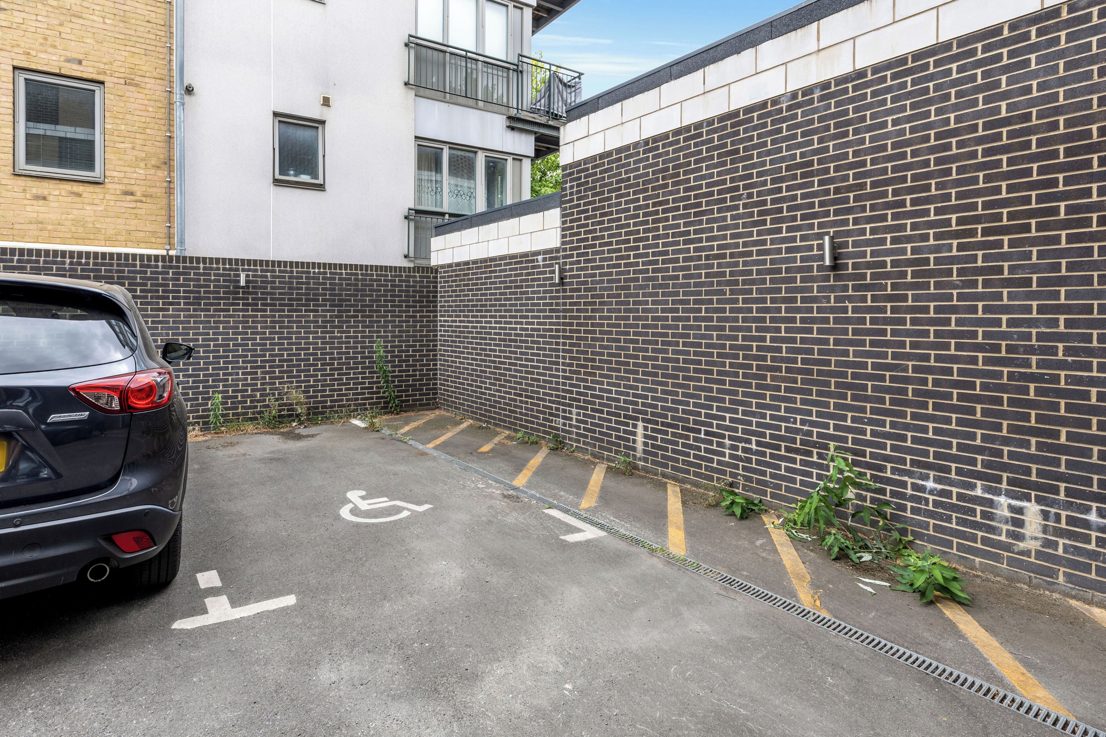 A parking area with a black car parked on the left, a wheelchair accessibility symbol on the ground, and a brick wall on the right.
