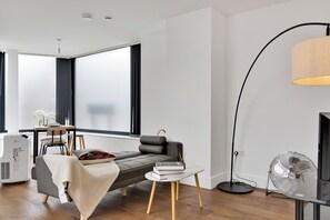 Een moderne woonkamer met een bank, een bijzettafeltje, een staande ventilator en een groot raam.