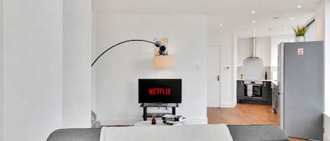 Un salon moderne avec un téléviseur à écran plat affichant le logo « NETFLIX », une petite table avec des livres, un plaid blanc et un luminaire au design incurvé.