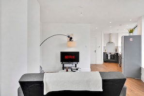 A modern living room with a flat-screen TV displaying 'NETFLIX,' a small table with books, a white throw blanket, and a curved light fixture. - Spacious Flat (ground floor) in Lambeth with gated parking (London)