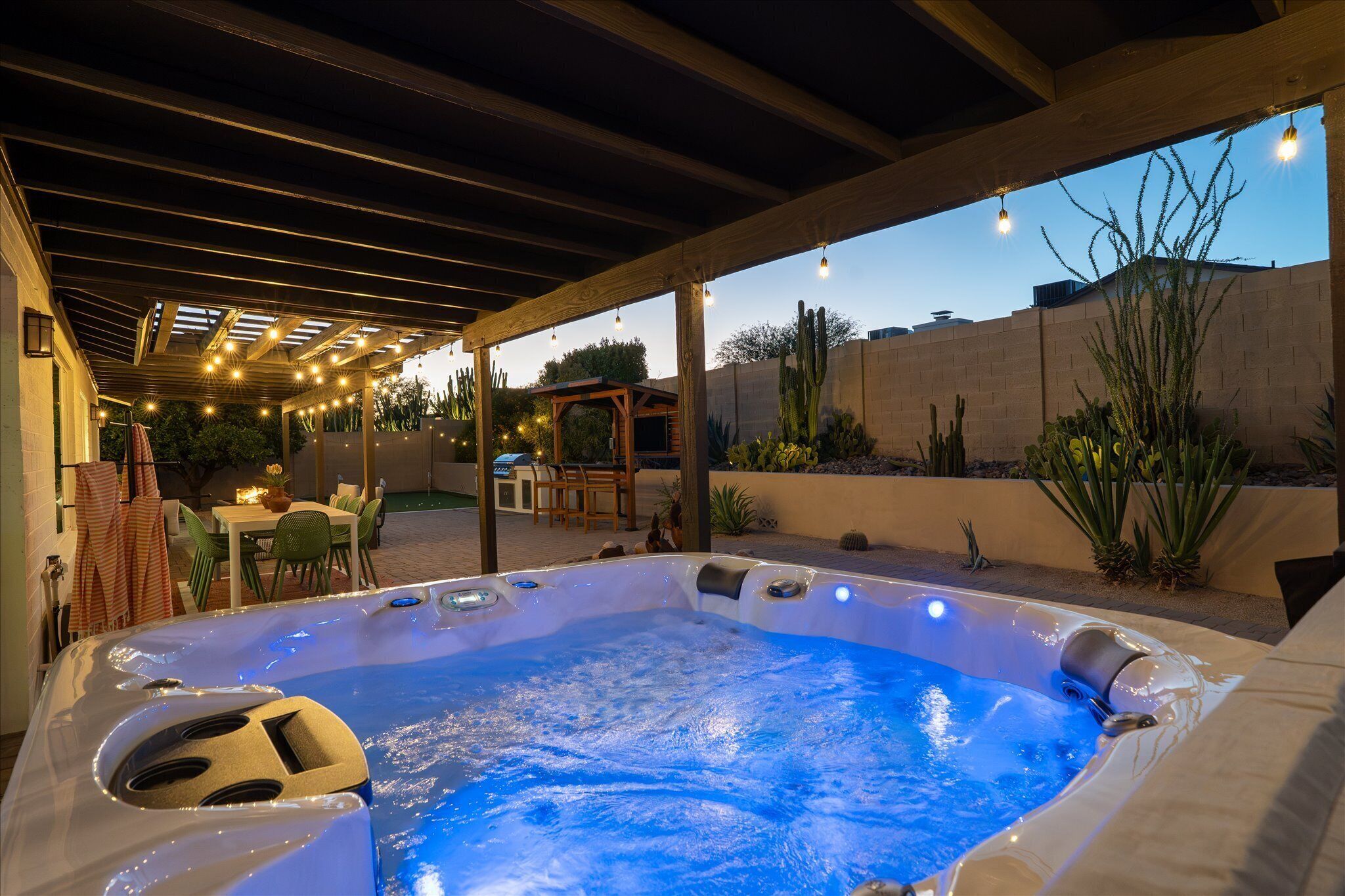 A hot tub under a wooden pergola with string lights, surrounded by a patio area with chairs and a table.