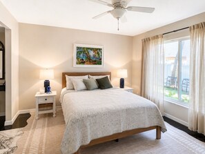 A bedroom with a bed, two bedside tables, a ceiling fan, a window with curtains, and a painting on the wall. - Coastal Retreat located on Manasota Key, Heated Pool, Beach & Bay Access (Englewood)