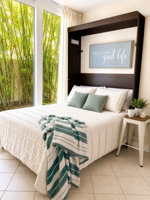 A bedroom with a bed, a bedside table with a plant, a framed quote on the wall, and a view of bamboo outside. - Coastal Retreat located on Manasota Key, Heated Pool, Beach & Bay Access (Englewood)