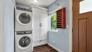 A laundry room with a front-loading washer and dryer, a white cabinet, a window with a view, and a wooden door. - Indigo Hideaway | Romantic + Fire pit (Nappanee)