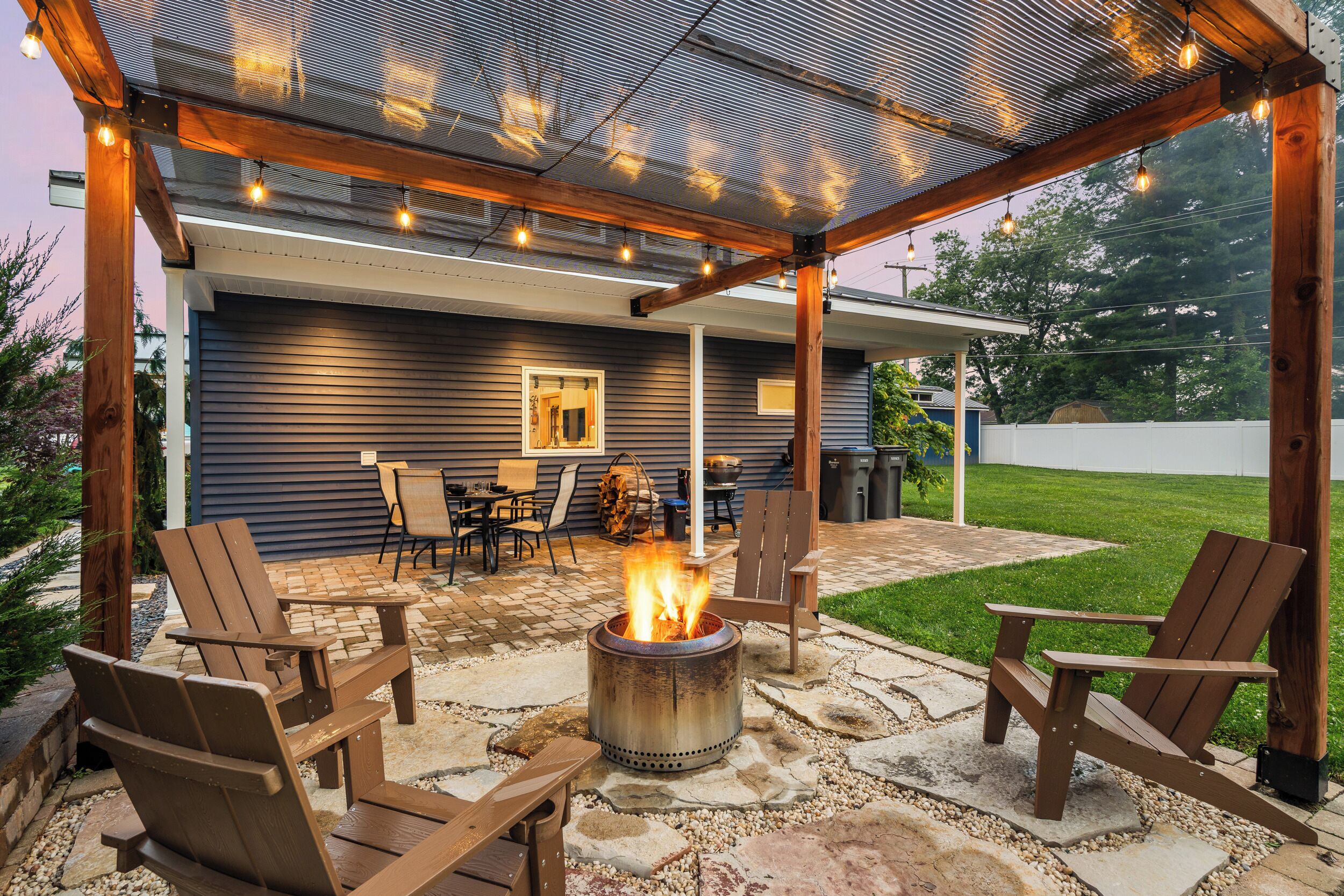 An outdoor patio with a fire pit, wooden chairs, and a covered seating area.