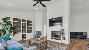 A modern living room with a sofa, a coffee table, a fireplace, a TV mounted on the wall, and a ceiling fan. - Remodeled 4BDR Home with Valley Views & Hot Tub (Draper)