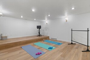 Ein Zimmer mit Holzboden, Trainingsmatten, Kurzhanteln, einem Fernseher auf einem Standfuß und einer schwarzen Hantelstange.