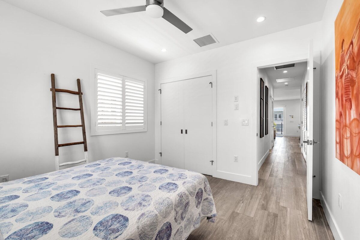 A bedroom with a bed, a ceiling fan, a window with blinds, a wardrobe, and a door leading to another room.