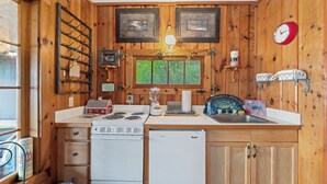 A compact kitchen with a stove, refrigerator, and sink. There are wooden walls and a window with a view.